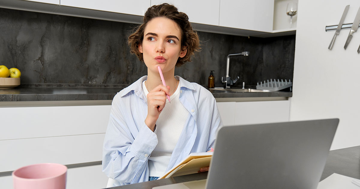 Ingyenes vagy fizetős online marketing tanfolyam? Döntés előtt Gondolkodó nő ül a laptopja mellett, kifelé nézve, azt mérlegelve, hogy ingyenes vagy fizetős online marketing képzést válasszon.