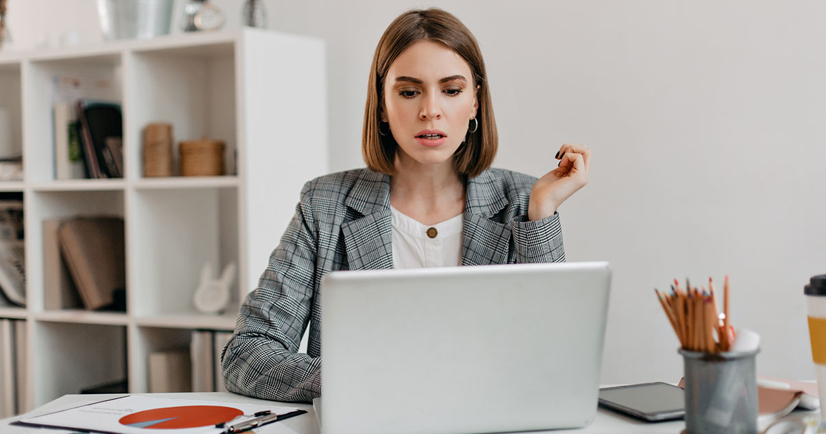 Nő laptop előtt ül asztalnál egy irodában, döntési helyzetet sugalló gondolkodó testtartással, hogy melyik online marketing képzést tanulja meg.